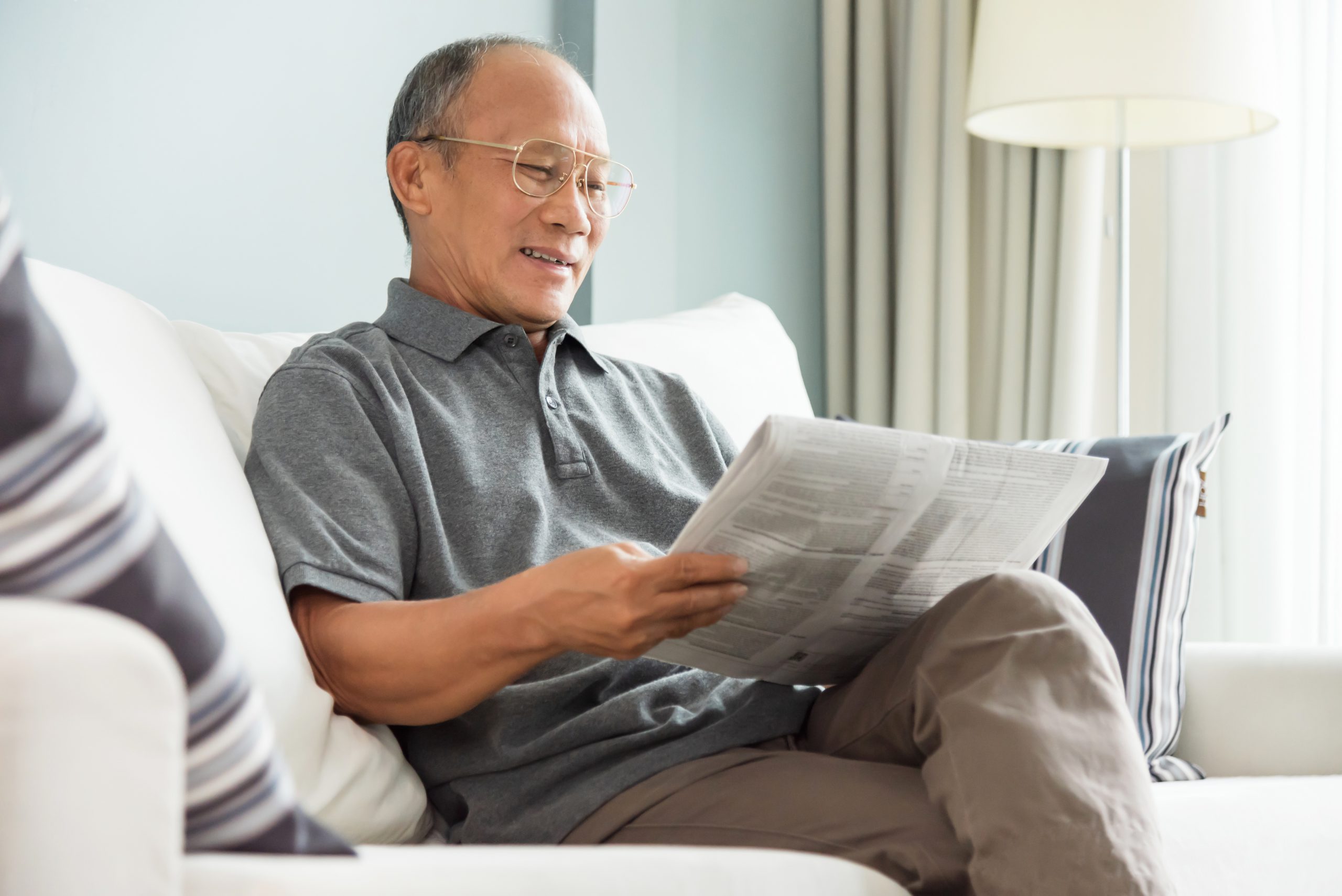 Happy Asian senior man sitting and reading newspaper at his house. Relaxing, Smiling, Retirement.
